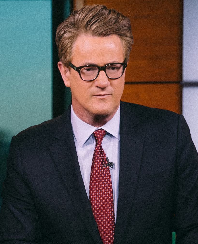 Joe Scarborough in a dark suit and red tie, studio headshot.