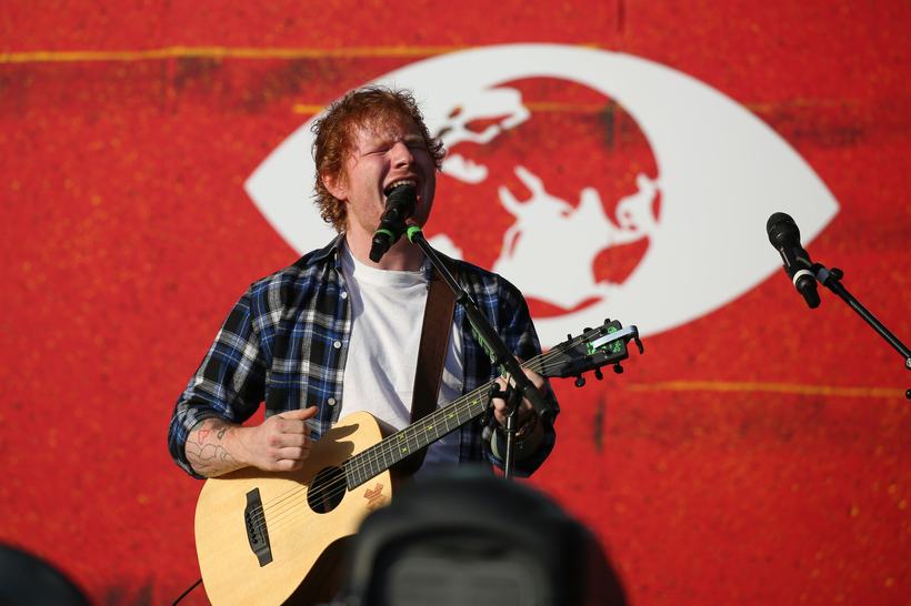 Ed Sheeran performing on an acoustic guitar in front of the red Global Citizen backdrop.