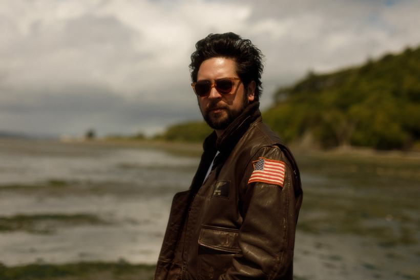 A man in sunglasses and a leather jacket on a grey shoreline.