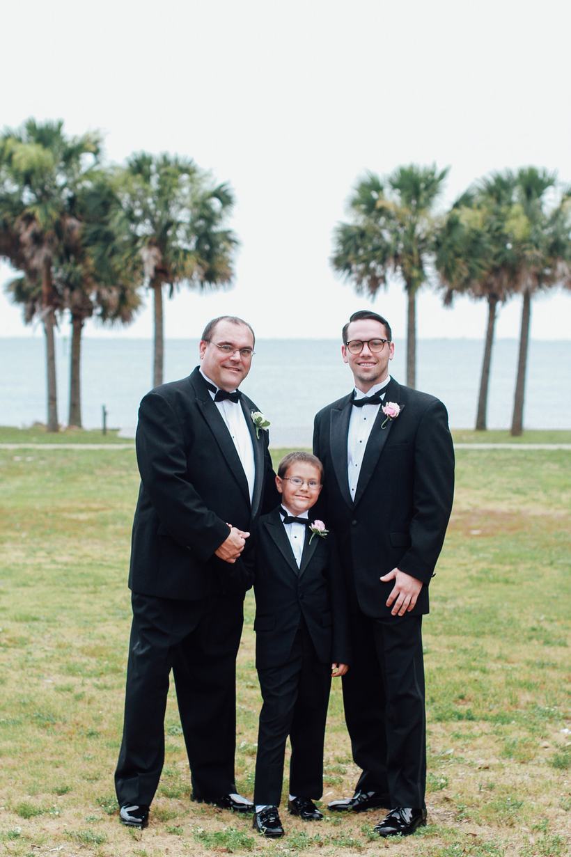 Three men in tuxedos standing on a lawn under palm trees.