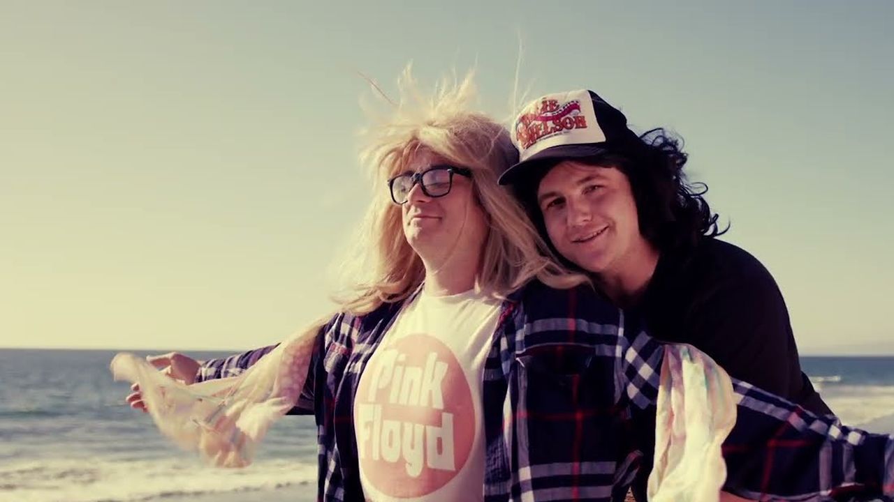 Two actors in wigs and band tees striking a pose on a beach at sunset, a still from the commercial.
