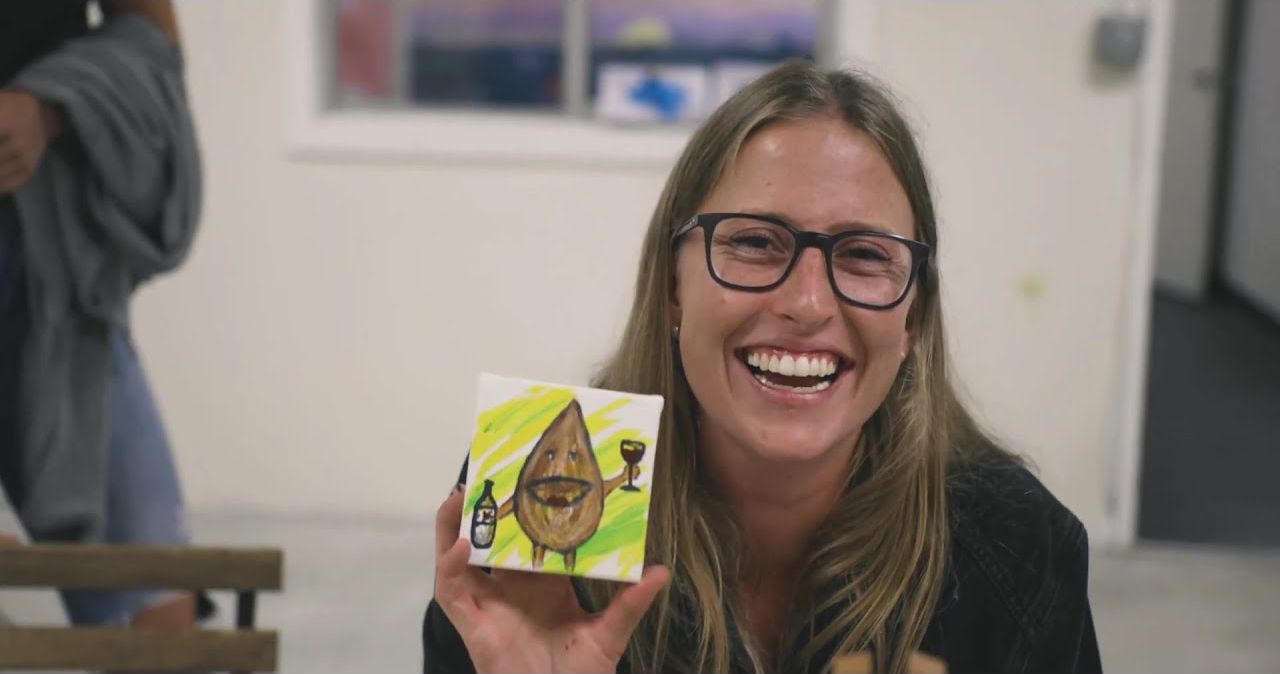 A guest laughing and holding up her finished painting at a paint night event.